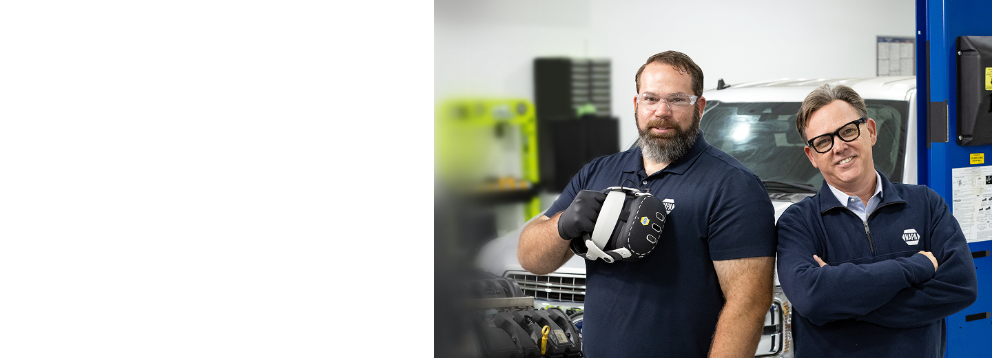 Two NAPA AutoCare representatives standing in an automotive training facility, with one holding a virtual reality headset and a vehicle in the background.