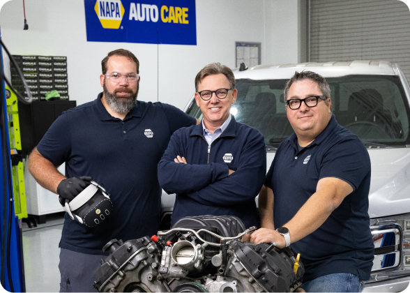 Three Xccelerator team members stand beside a vehicle engine in an automotive training workshop with a service vehicle in the background.